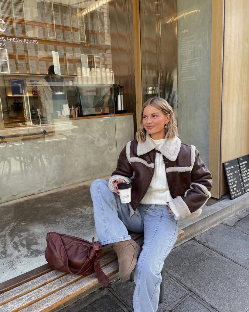 Woman in shearling jacket, light jeans, and Ugg boots with brown bag; stylish layered Ugg outfit.