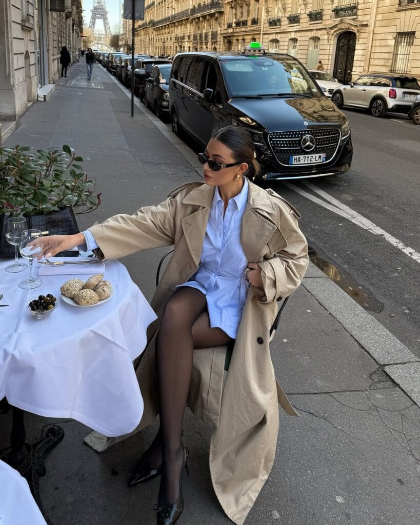 Woman in beige trench coat, blue shirt dress, and black heels at a Paris café table with Eiffel Tower view