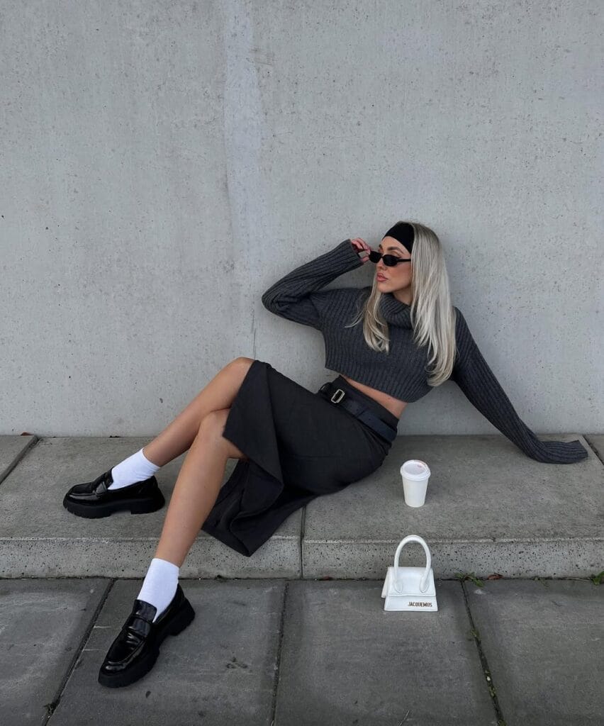 Woman in charcoal knit crop top, skirt, white socks, and black loafers sitting on sidewalk; loafers outfit with modern edge.