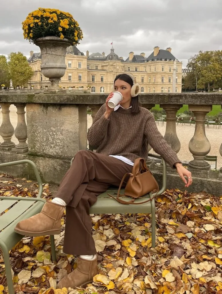 Woman in brown sweater and pants with Ugg boots and earmuffs, sitting outdoors in fall leaves.
