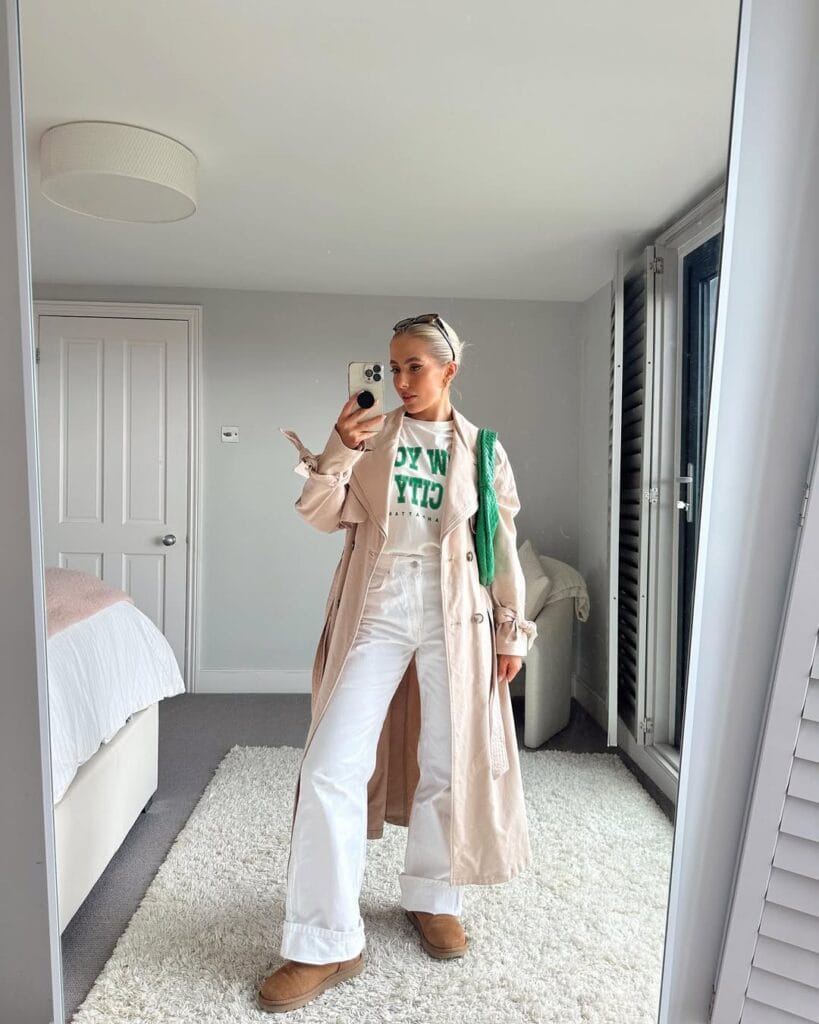 Woman in light trench coat, white jeans, graphic tee, and tan boots in a bedroom mirror selfie