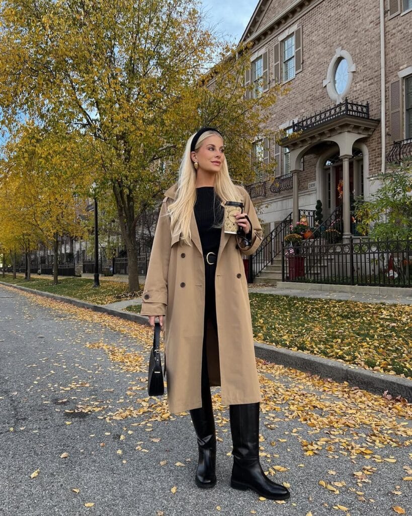 Woman in tan trench coat, black top, tall boots, and headband with coffee on a city street