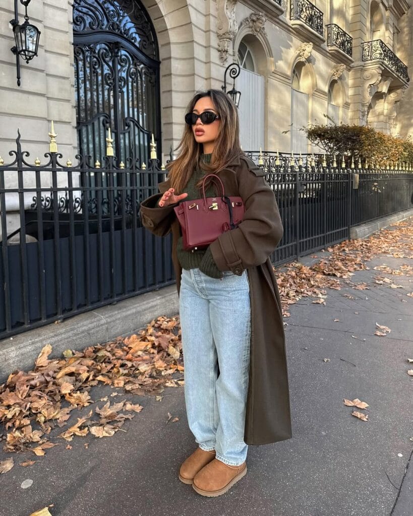 Woman in olive sweater, brown coat, jeans, and Ugg boots with burgundy bag; city chic Ugg outfit.