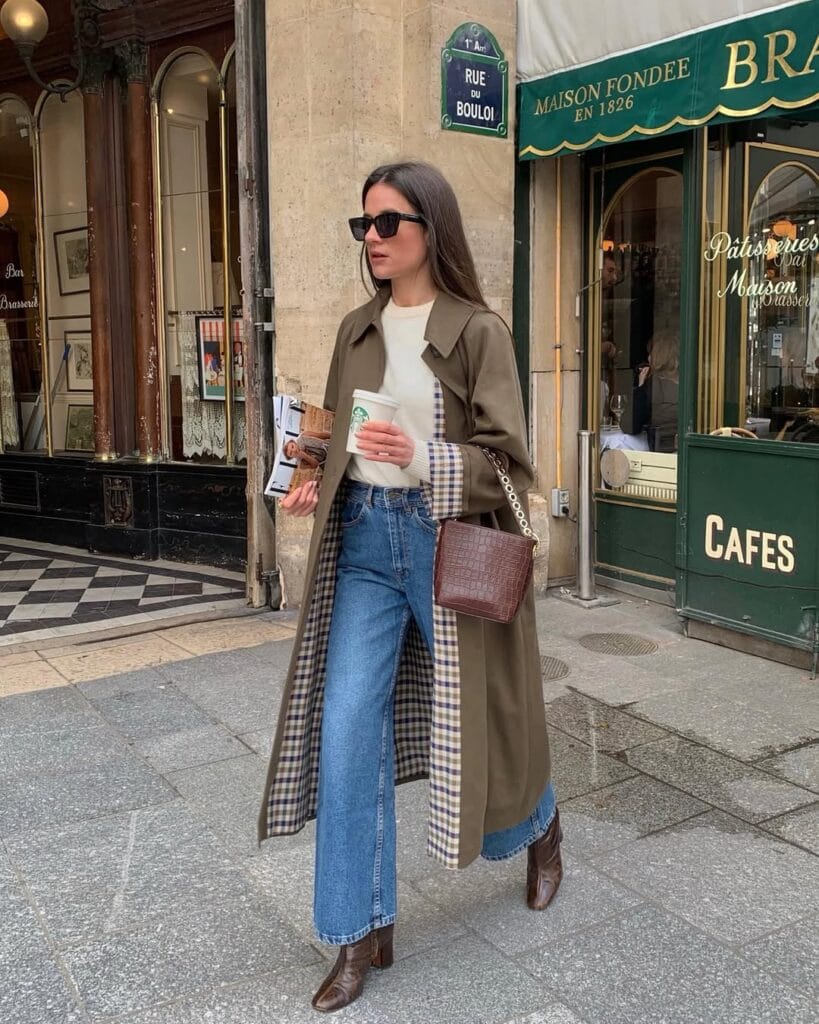 Woman in green trench coat, flared jeans, boots, and sunglasses walking past Paris café