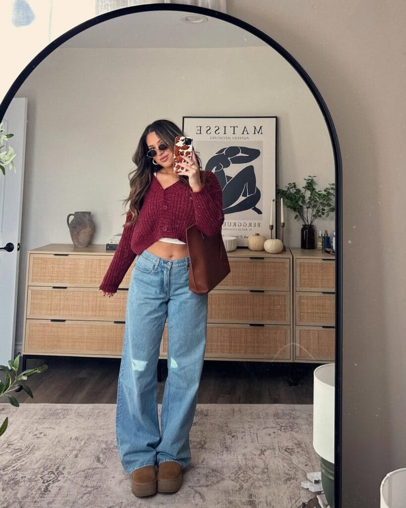 Woman in cropped red cardigan, wide jeans, and Ugg platform boots, taking mirror selfie; trendy Ugg outfit.