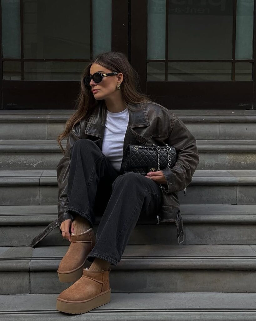 Woman in black leather jacket, wide-leg jeans, and Ugg platform boots, sitting on steps; edgy Ugg outfit idea.