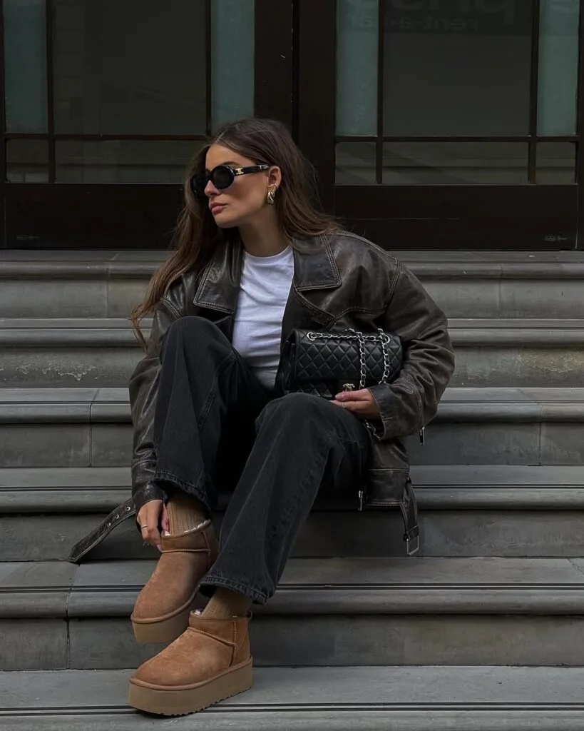 Woman in black leather jacket, wide-leg jeans, and Ugg platform boots, sitting on steps; edgy Ugg outfit idea.
