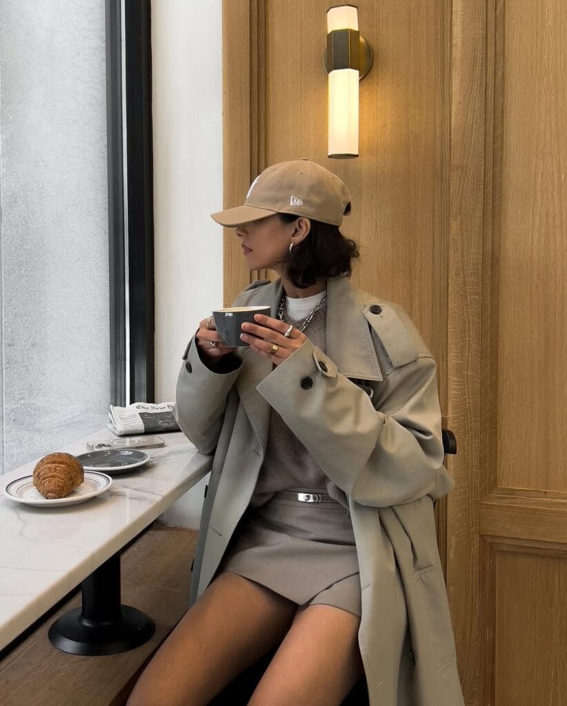 Woman in neutral trench coat, mini skirt, sweater, and baseball cap enjoying coffee at a window seat