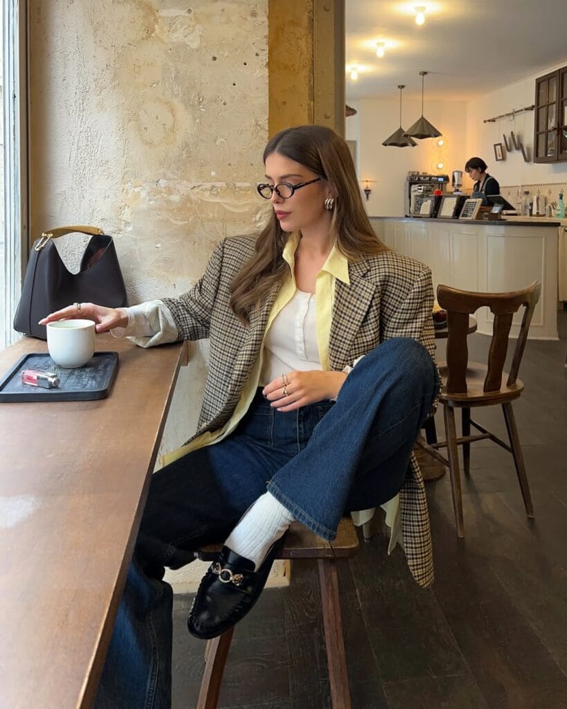 Woman in plaid blazer, jeans, black loafers, and glasses sitting at café; loafers outfit with academic flair.