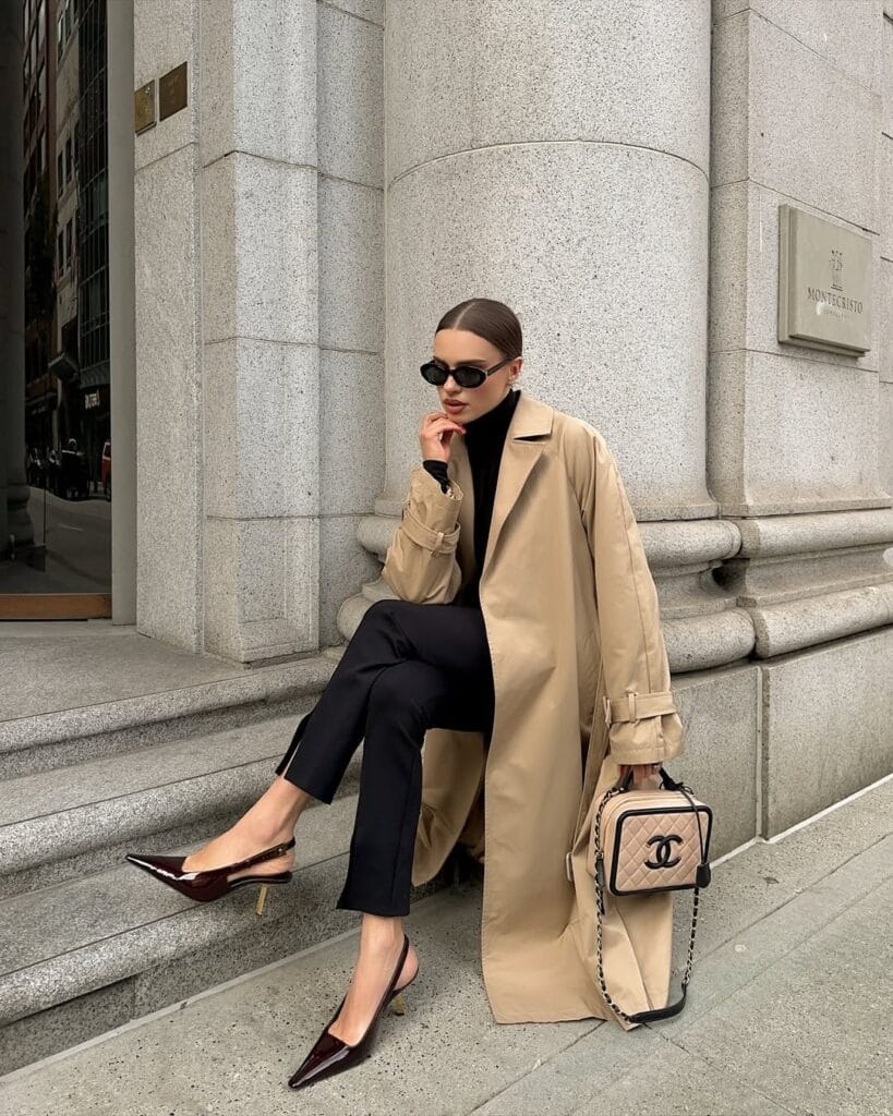 Woman in tan trench coat, black turtleneck, tailored pants, pointed heels, and Chanel bag sitting on steps